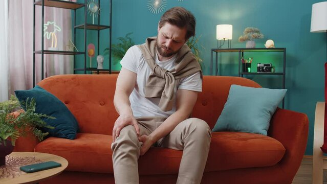 Young man suffer from painful severe knee joint ache, leg muscles, could not walk. Portrait of Caucasian guy massaging his knees illness cramps rheumatism at home room on sofa. Health care problem
