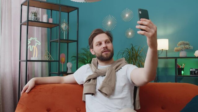 Happy man blogger taking selfie on smartphone, communicating video call online with subscribers, recording stories for social media. Portrait of Caucasian guy at home apartment living room on sofa