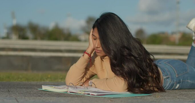 Young teenage Latina college student studying for an exam in an outdoor park with books