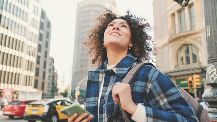 Happy girl using app on smartphone while traveling. Young smiling woman explores the streets, looks up