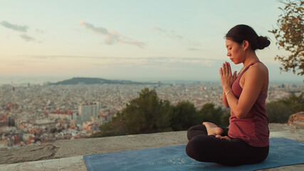 Fototapeta na wymiar Young woman in bodysuit practices yoga at dawn at viewpoint. Girl raises her hands up while sitting in lotus position