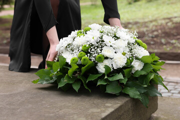 Woman with wreath of flowers near tombstone outdoors, closeup. Funeral attribute