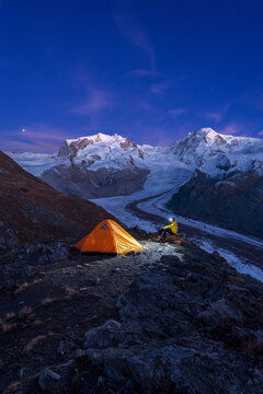 Hiker with head lamp sitting beside the tent in front of Monte Rosa massif, Gorner glacier (Gornergletscher) and Lyskamm peaks at dusk, Riffelsee, Zermatt, canton of Valais, Swiss Alps, Switzerland