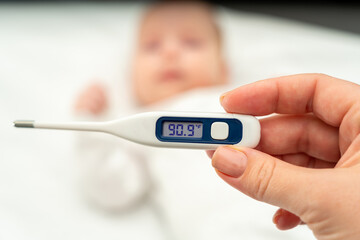 A womans hand holds a thermometer showing the temperature on the background of a blurred baby lying on the bed