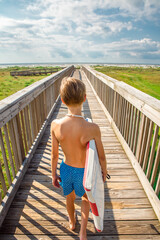 Young boy in swim trunks walking on a bridge toward the beach while carrying his boogie board