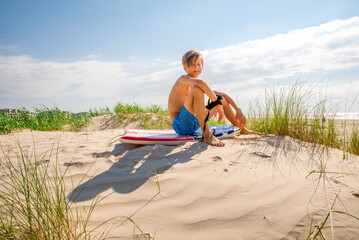 Happy young boy sitting on sand dune at the beach leashed to his boogie board