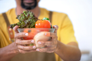 Man with black beard wearing an apron and mustard yellow t-shirt holds bowl of salad in hand on white background, selective focus