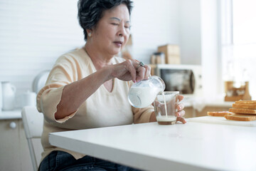 Asian grandmother pours milk into a cup and drinks in the morning, wellness and healthy lifestyle concept, selective focus