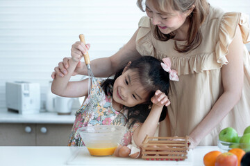Happy little girl helping her mother prepare breakfast in the kitchen, use a whisk to whisk the eggs in the bowl to make omelette, happy family concept