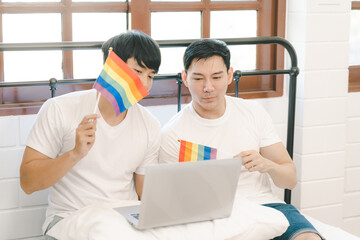 Happy engaged asian gay couple waving lgbt rainbow flag and VDO call to share happy moment at bedroom. happy married homosexual gay couple. Young Asia Gay couple feeling happy showing ring at home.