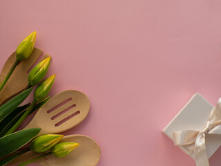 Yellow green tulips flowers with kitchen tools and white gift box on pink background with copy space. Women and Mothers Day greeting card. Birthday congratulations note. Utensil bouquet flat lay.