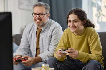 Teenage boy playing game console together with his dad while they sitting on sofa in front of TV in...