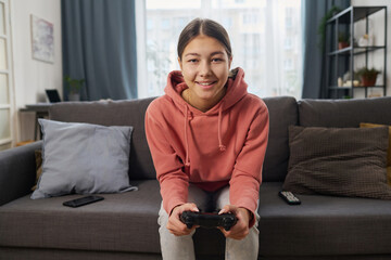 Portrait of smiling teenage girl looking at camera while sitting on sofa in the room and playing...