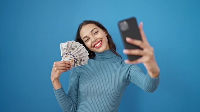 Young beautiful hispanic woman taking selfie holding dollars over isolated blue background