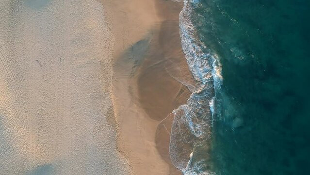 Aerial view of drone flying above sea, surf, and water crashing onto the sandy beach from a top angle