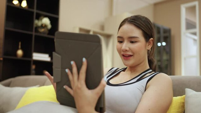 Asian woman in sportswear using digital tablet for online shopping or watching movie after workout exercise with online fitness training on laptop computer in living room. Home fitness gym concept.