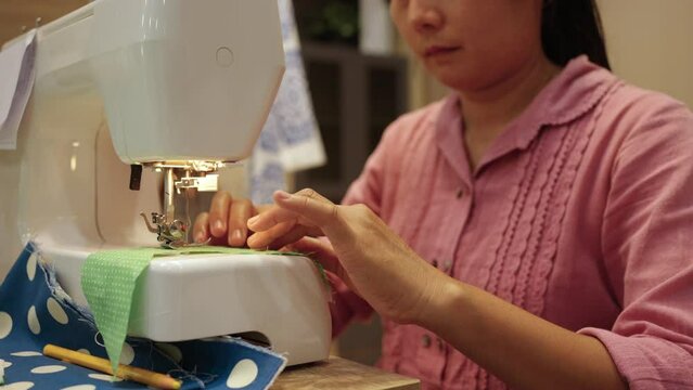 Mature Asian woman tailor stitching clothes on the sewing machine. Woman enjoy indoor activity lifestyle with handmade hobby making diy clothes and home decoration fabric in living room at home.