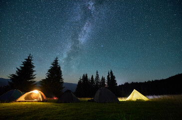 Night camping in mountains under starry sky. Tourist tents in campsite near burning campfire under beautiful sky full of stars with Milky way above forest. Concept of tourism and traveling.