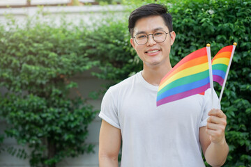 close up adult asian man raise rainbow flag (sign of lgbtq community) and smile with proudly at garden outside for show real personality and support marriage equality on pride month in June concept