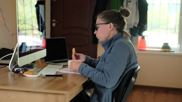 Girl drinks tea with cookies working at home. Woman in glasses work at home office, with personal computer and laptop lying on the table. Modern work culture and productivity.