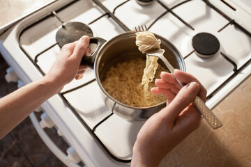 Hot instant pasta is cooked in a saucepan on the stove in the kitchen. The girl stirs the food in the bowl with a fork. Image for your creative decoration and design.