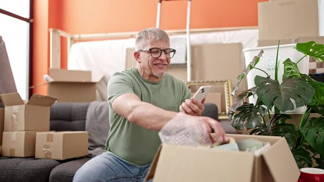 Middle age grey-haired man unpacking cardboard box make picture by smartphone at new home