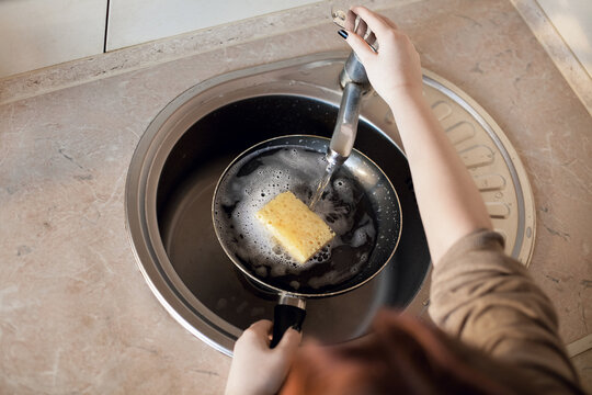 The girl washes a large dirty frying pan with a sponge and soap under running water under the tap. The plot is about housework and cleanliness in the kitchen.