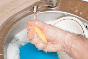 Fototapeta premium Washing dishes. A man's hand squeezes a soapy solution from a sponge, with a blue rag for washing dishes in the kitchen. An image about housework, apartment care and cleanliness.