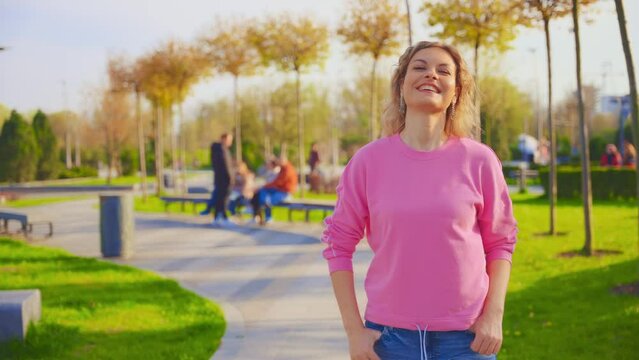 Portrait young beauty happy woman walking in city street blonde long hair fly in wind. Sexy girl fashion model smiling face enjoys park nature green grass trees sun light blue sky. pink shirt jeans 4k
