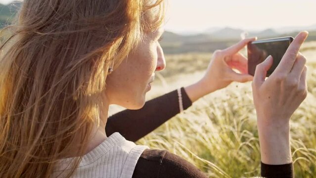 Side view slow motion woman filming nature with a smartphone in the sun glare sitting enjoying nature against the backdrop of a feather grass shining in the rays of a sunset