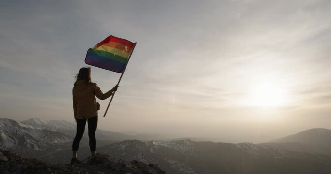 Successful silhouette woman winner waving LGBT pride flag on top of the mountain peak. liberal propaganda, political agenda. Cinematic background. Copy space