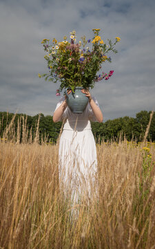 woman in white dress in wheat field holding vase with wild flowers 