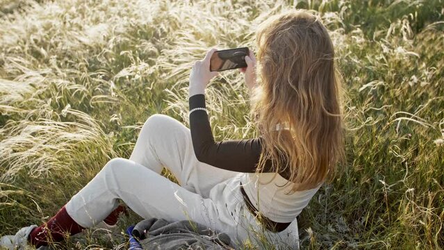 A modern woman sits enjoying nature against the backdrop of a feather grass shining in the rays of a sunny sunset and the wind blowing her hair. She films it all with her smartphone