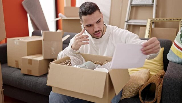 Young hispanic man unpacking cardboard box arguing by smartphone at new home