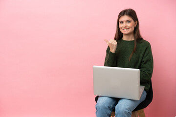 Naklejka premium Young caucasian woman sitting on a chair with her laptop isolated on pink background pointing to the side to present a product