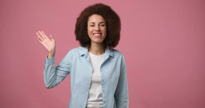 Happy friendly young african american woman waves palm in hello gesture welcomes someone with hospitable expression expresses positive emotions standing over pink background