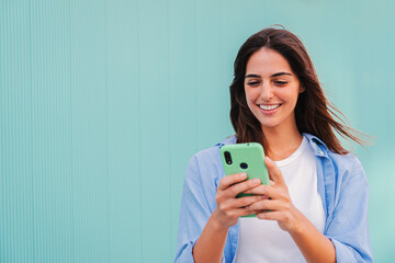 Smiling happy teenage woman using her smartphone and chatting online ,or browsing on internet at blue teal wall background. Young caucasian college student lady watching funny videos with mobile phone - obrazy, fototapety, plakaty