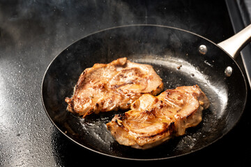 Pork steak is fried in hot splattering and steaming oil in a black iron skillet on the stove top...