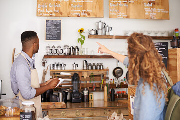 Woman, cafe and pointing at menu on wall with barista, notes and decision for good customer...