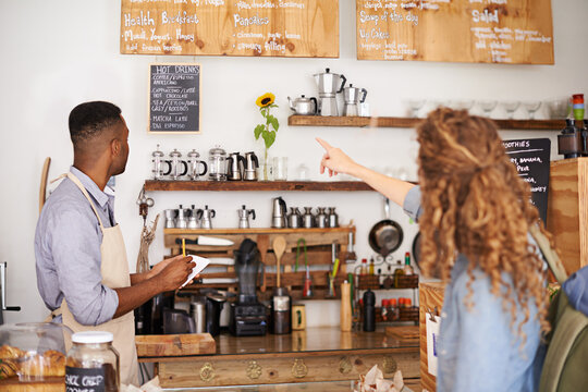 Woman, cafe and pointing at menu on wall with barista, notes and decision for good customer experience. Male waiter writing, lady and talking with choice, thinking and order special from bakery shop