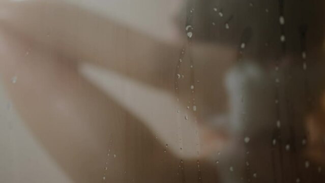 Close-up of a European-looking woman taking a rain shower. Water flies falls. Warm water in the shower. The girl washes slowly, applying shower gel on her naked body
