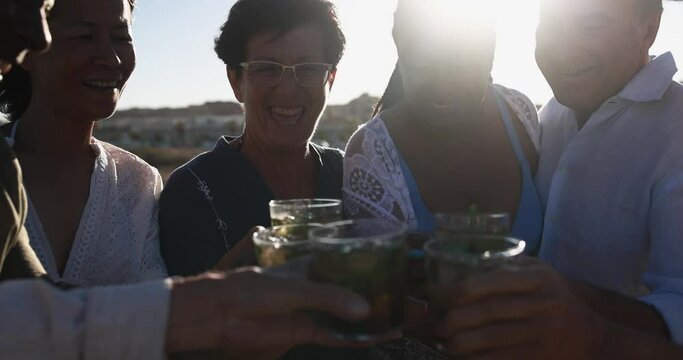 Senior multiracial friends cheering with alcohol free mojitos on the beach at sunset - Mature people having fun during summer vacation