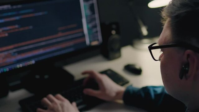 A young middle-aged programmer works remotely in his city apartment at night. IT specialist sits in front of the monitor of his laptop computer. Web programming writing website code, working remotely
