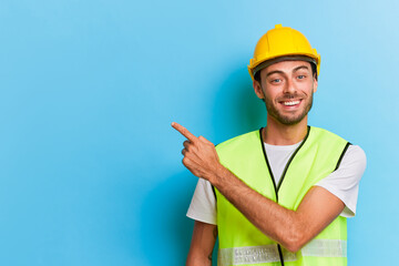 Cheerful bearded worker in yellow safety helmet and yellow vest smiles happily, points his finger out empty space for your ads, professional people concept, copy space