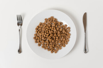 Dry animal food in a white ceramic plate served with knife and fork on a beige neutral studio background. Cat or dog food. Creative funny image for ad of pet products. Top view, flat lay, copy space