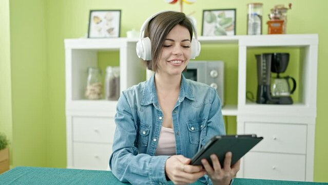 Young caucasian woman using touchpad and headphones sitting on table at home