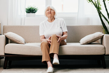Elderly woman severe pain in her leg sitting on the couch, health problems in old age, poor quality of life. Grandmother with gray hair holds on to her sore knee, problems with joints and ligaments.  - obrazy, fototapety, plakaty