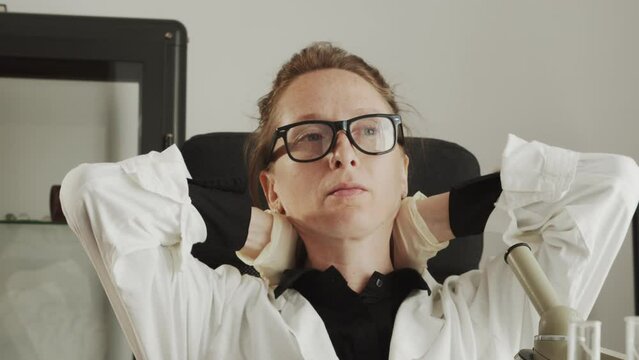 A young adult woman doctor in glasses and a white coat is tired and kneads her stiff neck while examining with a microscope. Scientific activity and hobby