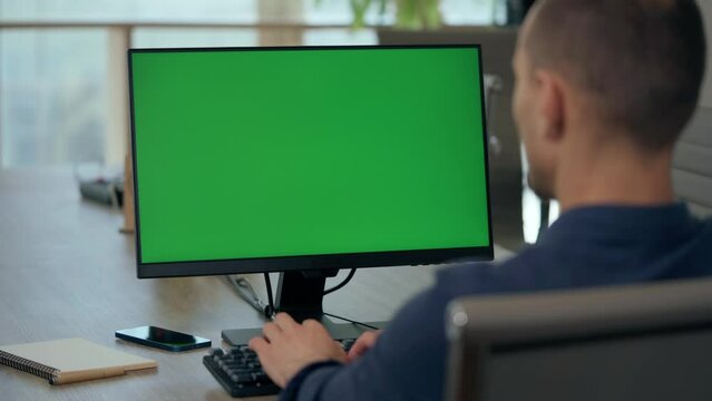 Over the Shoulder: Young Man Sitting at His Desk in Office Uses Desktop Computer with Green Mock-up Screen. Close up Green Screen Mock Up Display