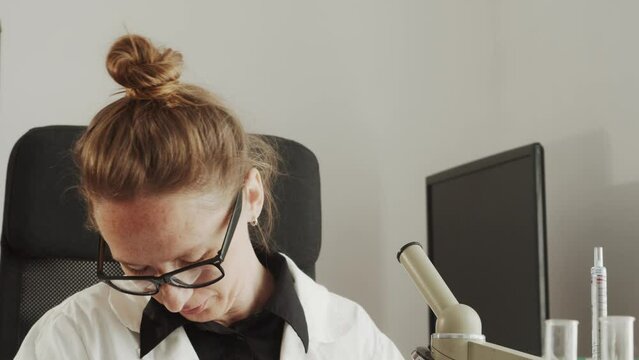 A young adult woman doctor in glasses is tired and kneads her stiff neck while doing entomology research activities in a laboratory in a white coat. Scientific activity and hobby